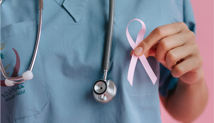 Attēls ar sievieti ārsta formā, stetoskopu ap kaklu, turot rokā rozā lentīti  - simbolu, kas demonstrē izpratni un atbalstu krūts vēža skartajiem cilvēkiem